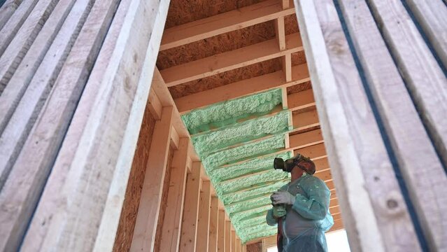 Male builder insulating wooden frame house. Man worker spraying polyurethane foam inside of future cottage, using plural component gun. Construction and insulation concept.