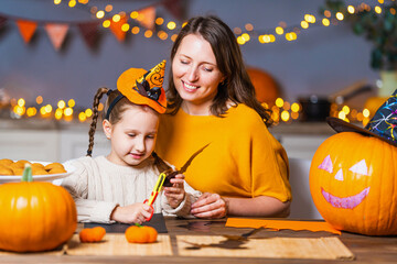 Fototapeta premium loving family, mom and daughter, cut out paper figures, making decorations for house for Halloween, at wooden table in kitchen. woman and girl draw bats and pumpkins, lights of garlands in background