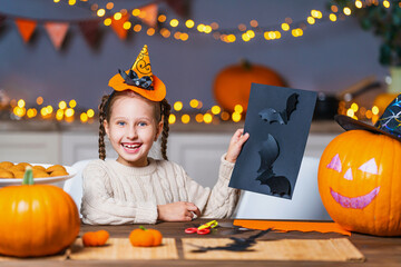 cute little girl with a hat on her head, sitting at a table in the kitchen at home, carves bats out...