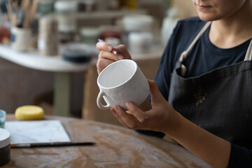 Craftswoman creates new masterpiece in work studio making sketch of future pattern on white mug....