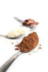 From above of metal spoons with cocoa, coconut flakes, lavender, rose petals