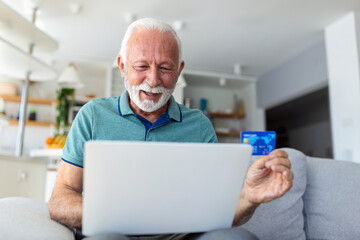 Fototapeta premium Mature man using laptop, holding plastic credit or debit card, senior grey haired customer making secure internet payment, shopping or browsing online banking service, entering information