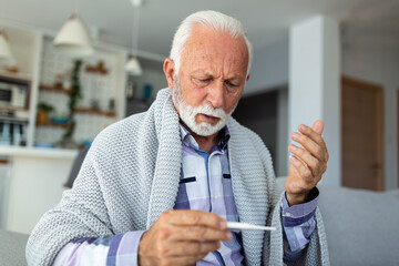 Sick elderly man checking his temperature suffering from seasonal flu or cold lying on sofa...