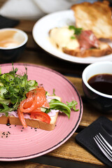 classic avocado toast with cucumber, salmon, microgreens, arugula and sauce on bread in a plate. cappuccino and filter coffee in the background. close-up