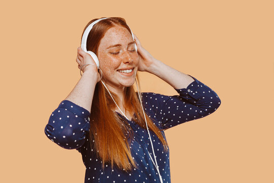 Young Beautiful Woman With Ginger Hair Red Head, Freckles And Blue Eyes, Wears Earphones, Listens Music With Eyes Closed At Studio Over Beige Background. Technology Concept.