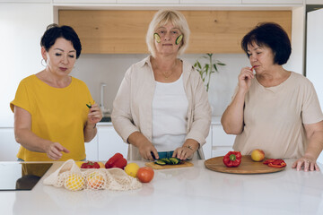 Portrait of a beautiful senior woman with cucumber slices on her cheeks. A group of friends preparing food together in a kitchen.