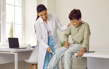 Friendly doctor examining little child. Professional neurologist uses hammer to check knee reflex of happy school boy sitting on medical couch in exam room. Children, health checkup, neurology concept - obrazy, fototapety, plakaty