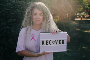 Blond Caucasian woman with curly hair recovering after cancer standing outdoors. Pink ribbon and...