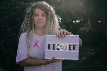 Blond Caucasian woman with curly hair recovering after cancer standing outdoors. Pink ribbon and...
