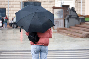 Rain in city, slim woman with umbrella wearing jeans and jacket standing on а street on crosswalk background. Rainy weather in autumn