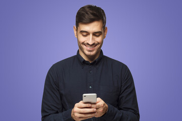 Young smiling businessman in shirt reading sms, using smartphone on purple background