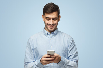 Young smiling business man in blue shirt reading sms, using phone on blue background
