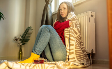 A woman wrapped in a plaid is sitting on the floor and warming herself near a warm radiator. Crisis of energy resources.