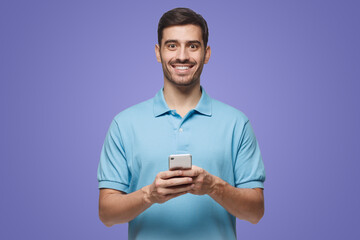 Young man standing on purple background with smartphone, looking at camera and smiling