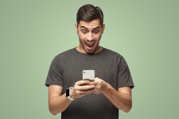 Young excited man looking at phone with surprise, isolated on green background