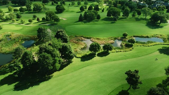 Aerial drone shot over golf club covered by lush green vegetation and small ponds at daytime.