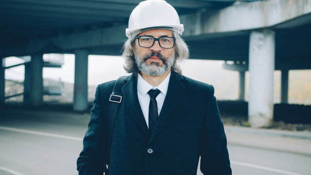 Portrait of respectful businessman standing in industrial zone outdoors wearing safety helmet and stylish suit and looking at camera with serious face