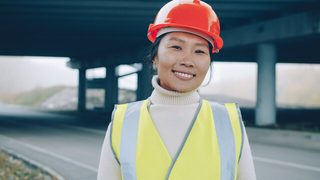 Asian forewoman wearing vest and helmet is standing outside in construction site smiling enjoying building career. People and occupation concept.