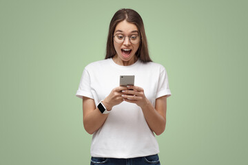 Naklejka premium Portrait of young woman in white t-shirt, holding phone, looking schocked and surprised