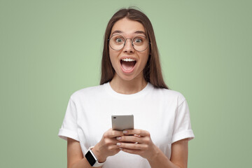 Close-up of girl in white t-shirt, holding phone, looking surprised and shocked, isolated on green