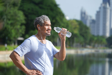 senior man with grey hair and beard wearing headphones for music listening is drinking water after workout in the city park,concept elderly people lifestyle, exercise, body,water substitute,health 