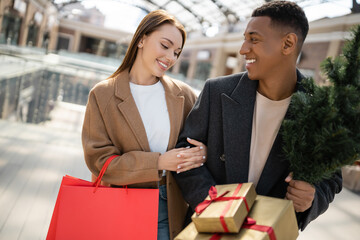 stylish african american man with small christmas tree and gift boxes looking at pleased girlfriend with shopping bags.