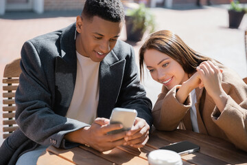 cheerful multiethnic couple looking at mobile phone on cafe terrace.