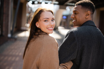 pretty and happy woman looking at camera near african american boyfriend on blurred street.