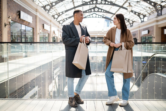 full length of interracial couple in coats and jeans holding shopping bags and looking at each other near mall.