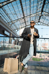 full length of african american man with coffee to go and smartphone standing near shopping bags and mall.