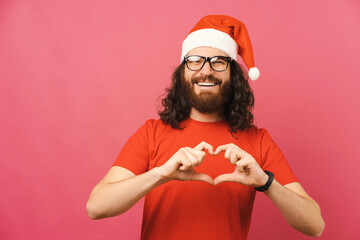 Handsome bearded man wearing glasses and Christmas hat shows heart gesture.