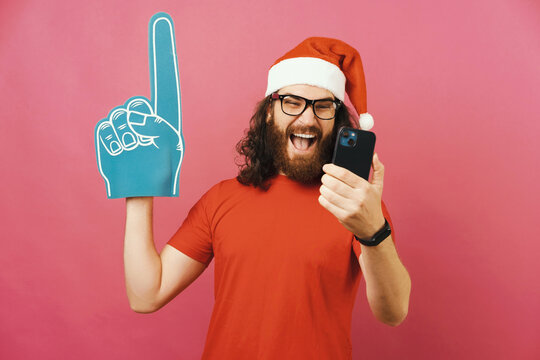 Cheerful man watches Christmas football match on phone while wearing fan glove.