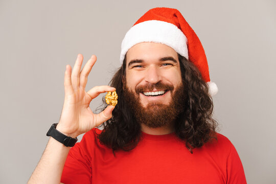 Healthy bearded man wearing a Christmas hat is holding a walnut near his face.