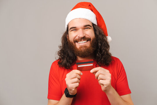 Content bearded man wearing Christmas hat is holding tight a red credit card.