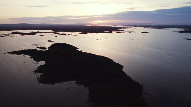 Beautiful aerial flying over gigantic irish lough (lake) corrib with many small island silhouettes in the water and more in the distance. Flying forward during blue hour golden evening sun reflecting.