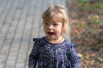 Portrait of a little blonde girl with an open mouth against the background of paving slabs and autumn leaves.