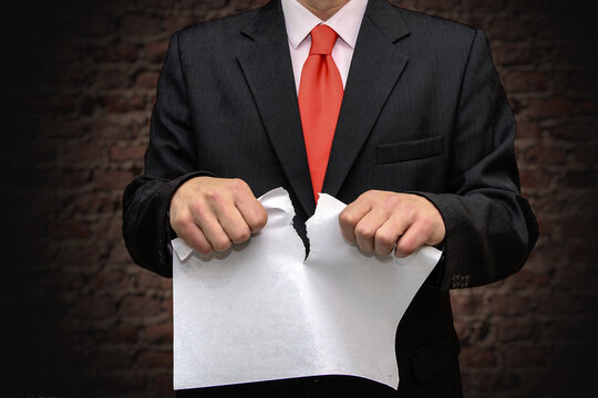 A Man In A Suit Tears Up A Piece Of Paper Against A Brick Wall. Concept: Termination Of The Contract, Invalid Document, Dismissal From Work.