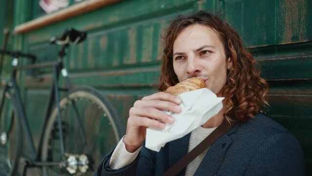 Happy curly haired man wearing jacket eating croissant in cafe outdoors