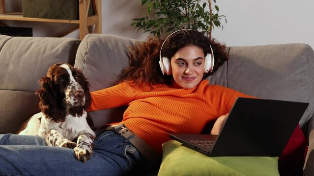 Young woman is using laptop and lying with English springer spaniel dog on bed at home. Beautiful girl hugging with her beloved pet and looking at computer screen with smile. Domestic animal