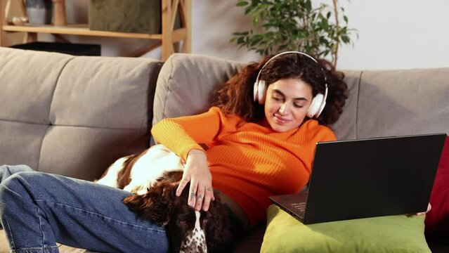 Young woman is using laptop and lying with English springer spaniel dog on bed at home. Beautiful girl hugging with her beloved pet and looking at computer screen with smile. Domestic animal