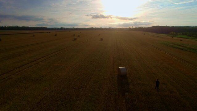 Lone man with a backpack travels on an adventure through a field of straw bales, to a dramatic sunset on the horizon. Distant aerial view