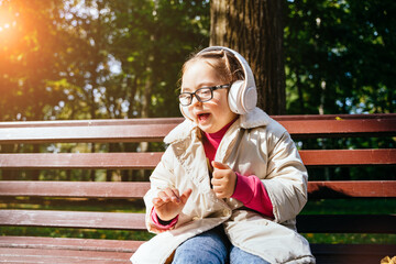 Portrait of cute lovely little girl listening music with headphones sitting on the bench at park...
