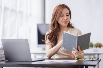 Fototapeta premium Asian Businesswoman holding notes and working on a laptop computer in the workplace, doing document planning analyzing the financial report, business plan investment, finance analysis concept.