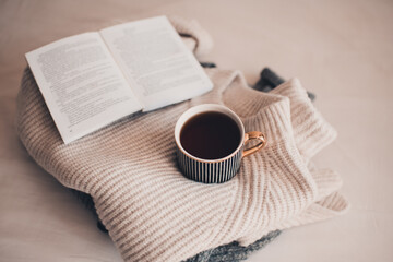 Obraz na płótnie Canvas Cup of coffee stay on stack of knitted textile sweater in bed with paper open book. Winter cozy season.