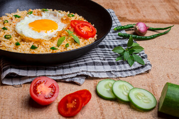 Fried rice served with fried egg, sliced ​​tomato, green chili and cucumber in a black skillet.