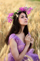 Young brunette woman in violet fairytale dress and pink flower crown sitting in wheat field....