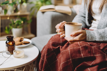 Cozy woman legs in knitted winter warm sweater and checkered plaid drinking hot cocoa or coffee in...