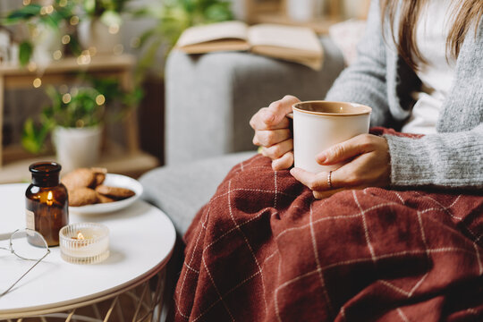 Cozy woman legs in knitted winter warm sweater and checkered plaid drinking hot cocoa or coffee in mug, sitting on couch at home. Autumn vibes with candle, decor, cookies and indoor plants