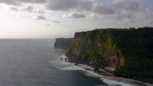 Silhouette of majestic Uluwatu cliffs standing tall beside coastline of beach and low tides moving towards them.