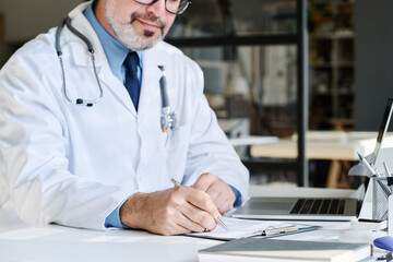 Close-up of mature doctor in white coat sitting at table at office and filling medical card of patient